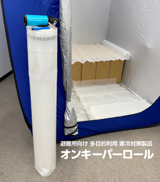 避難用の屋内テントの床と、テント内に設置された段ボールベッドの上に半透明のクッションシートが敷かれており、テントの外には巻かれたシートが立てて置かれている。